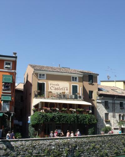 A street-level view of colorful buildings along a waterfront promenade in Sirmione, featuring the "Piccolo Castello" restaurant with outdoor seating and people walking nearby.