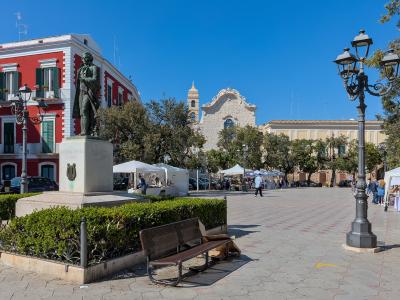 A wide public square featuring a bronze statue of a man on a stone pedestal, a red building with green shutters to the left, a church with a bell tower in the background, and market stalls set up in the distance.