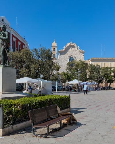 A wide public square featuring a bronze statue of a man on a stone pedestal, a red building with green shutters to the left, a church with a bell tower in the background, and market stalls set up in the distance.