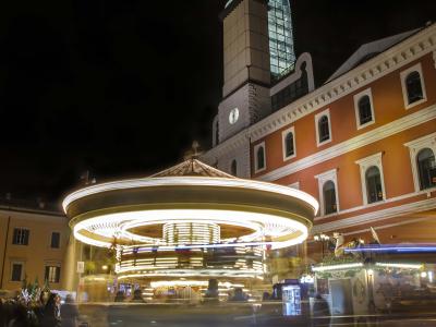A nighttime photograph of a historic Italian piazza featuring a prominent two-tiered stone fountain with lion sculptures, flanked by medieval-style buildings with arched loggias and a tall bell tower on the right, under a deep blue twilight sky.
