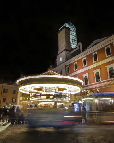 A nighttime photograph of a historic Italian piazza featuring a prominent two-tiered stone fountain with lion sculptures, flanked by medieval-style buildings with arched loggias and a tall bell tower on the right, under a deep blue twilight sky.