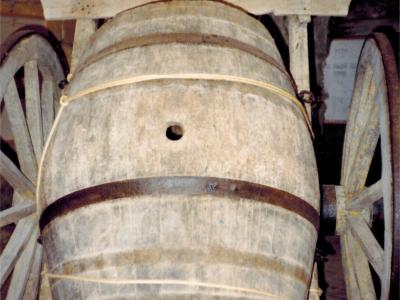 A weathered wooden barrel mounted on a rustic two-wheeled cart, displayed indoors with dim lighting and a dark background, likely within a museum setting.