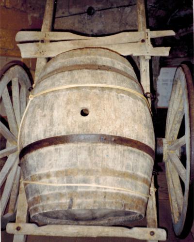 A weathered wooden barrel mounted on a rustic two-wheeled cart, displayed indoors with dim lighting and a dark background, likely within a museum setting.