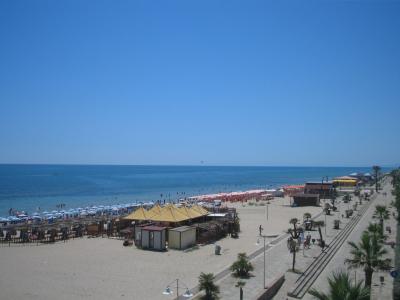 A wide sandy beach lined with rows of colorful umbrellas and sunbathers, featuring a paved promenade with palm trees and small beachside structures under a clear blue sky.