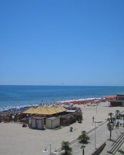 A wide sandy beach lined with rows of colorful umbrellas and sunbathers, featuring a paved promenade with palm trees and small beachside structures under a clear blue sky.