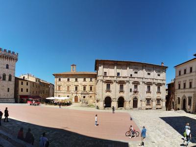A wide-angle photograph of a large, sunlit Italian piazza featuring a prominent medieval-style palace with a tall crenellated tower on the left, flanked by other Renaissance-era buildings, with people walking and sitting on the paved ground under