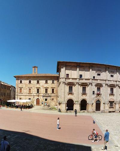 A wide-angle photograph of a large, sunlit Italian piazza featuring a prominent medieval-style palace with a tall crenellated tower on the left, flanked by other Renaissance-era buildings, with people walking and sitting on the paved ground under
