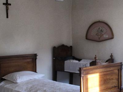 The image shows a sparsely furnished bedroom interior with two wooden beds, a hanging oil lamp, a crucifix on the wall, and a decorative fan, consistent with a historical museum room.