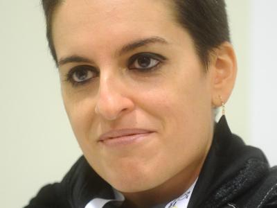 A close-up portrait of a woman with short dark hair wearing a black costume and a white lanyard with the text "SCUOLA DEL FUMETTO" visible.