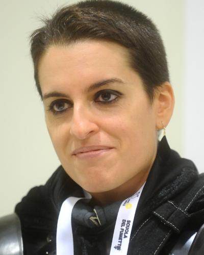 A close-up portrait of a woman with short dark hair wearing a black costume and a white lanyard with the text "SCUOLA DEL FUMETTO" visible.