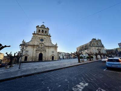 The image shows a large, ornate Baroque-style church facade with a central clock and bell tower, situated in a paved public square lined with pruned trees and street lamps, with another historic building visible to the right and modern cars parked