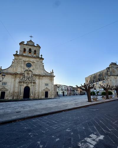 The image shows a large, ornate Baroque-style church facade with a central clock and bell tower, situated in a paved public square lined with pruned trees and street lamps, with another historic building visible to the right and modern cars parked