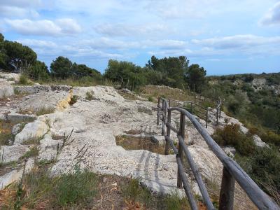 A rocky, sunlit landscape with a wooden railing winding along a path, surrounded by Mediterranean vegetation including pine trees and shrubs under a partly cloudy sky; no visible signage or structures identifying the location.