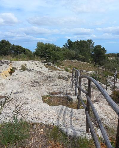 A rocky, sunlit landscape with a wooden railing winding along a path, surrounded by Mediterranean vegetation including pine trees and shrubs under a partly cloudy sky; no visible signage or structures identifying the location.