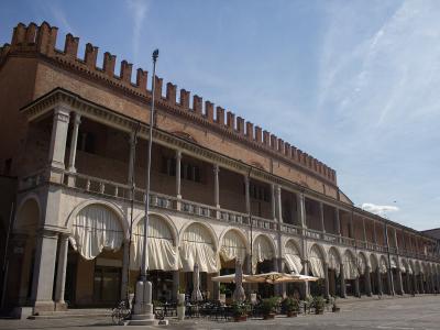 A long, medieval-style brick building with a crenellated roofline and a ground-floor loggia featuring stone columns and white draped awnings, situated in a paved public square under a blue sky.