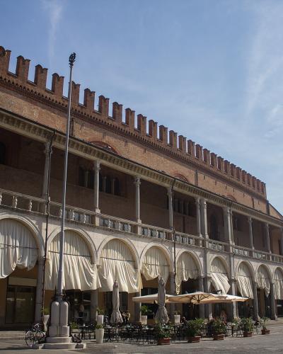 A long, medieval-style brick building with a crenellated roofline and a ground-floor loggia featuring stone columns and white draped awnings, situated in a paved public square under a blue sky.