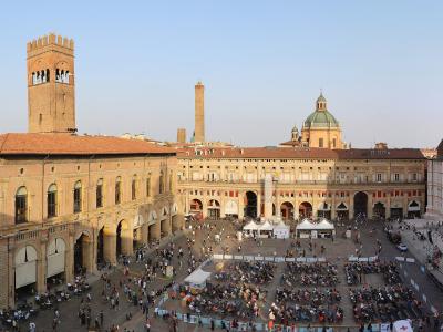 A high-angle panoramic view of a large, crowded medieval European square featuring the Palazzo Comunale with its prominent tower on the left, the Basilica di San Petronio on the right, and the Asinelli Tower in the background.
