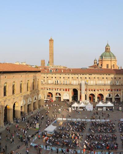 A high-angle panoramic view of a large, crowded medieval European square featuring the Palazzo Comunale with its prominent tower on the left, the Basilica di San Petronio on the right, and the Asinelli Tower in the background.