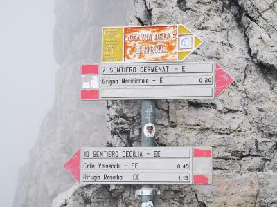 A close-up of a metal hiking trail signpost mounted on a rocky cliff face, featuring directional arrows for "ALTA VIA DELLE GRIGNE," "SENTIERO CERMENATI," and "SENTIERO CECILIA" with distances to destinations like Grigna Meridionale and Rifugio Ro