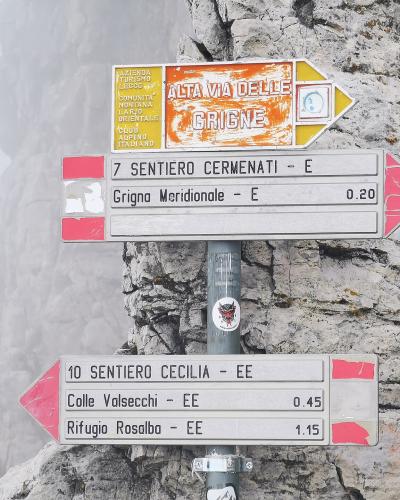 A close-up of a metal hiking trail signpost mounted on a rocky cliff face, featuring directional arrows for "ALTA VIA DELLE GRIGNE," "SENTIERO CERMENATI," and "SENTIERO CECILIA" with distances to destinations like Grigna Meridionale and Rifugio Ro