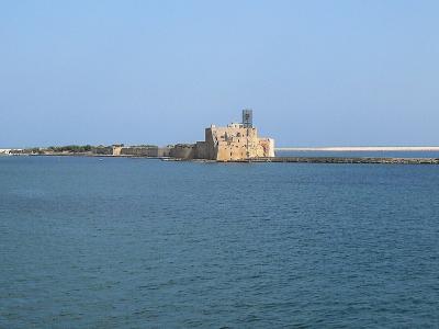 A wide-angle view of the Castello di Brindisi (Brindisi Castle) situated on a breakwater extending into the sea, with a long stone mole and calm blue water under a clear sky.