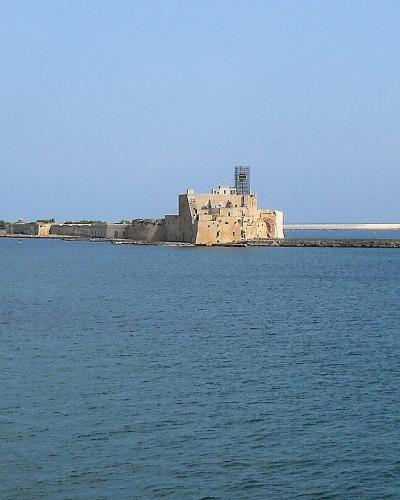 A wide-angle view of the Castello di Brindisi (Brindisi Castle) situated on a breakwater extending into the sea, with a long stone mole and calm blue water under a clear sky.