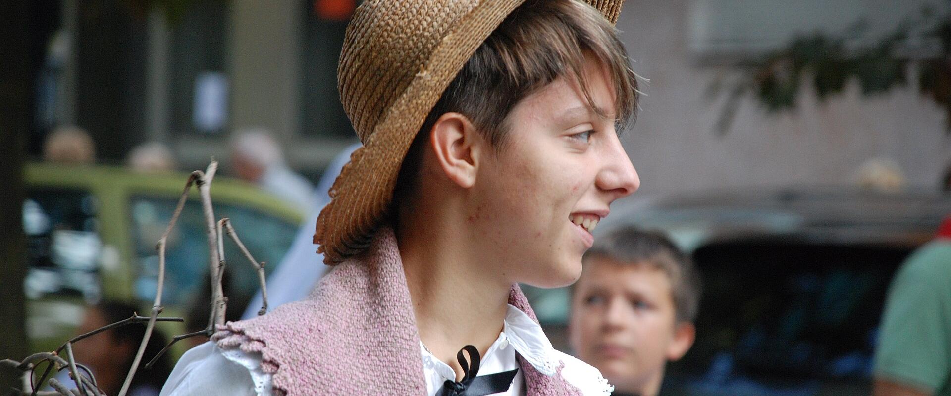 A young person in profile wearing a straw hat, white blouse, and pink knitted shawl, holding bare branches, with another child in similar attire behind them in an outdoor street setting.