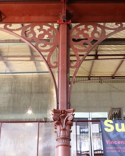 The image shows an interior view of a historic market hall, featuring ornate red cast-iron columns with Corinthian-style capitals and decorative scrollwork supporting a glass-and-metal roof structure; signage in the lower right includes Italian te