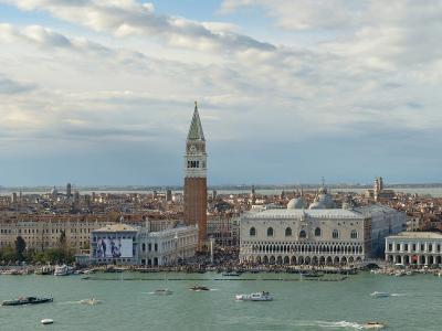 A panoramic view of the Venetian lagoon and skyline, prominently featuring St. Mark’s Campanile (bell tower), the Doge’s Palace, and the waterfront buildings of Piazza San Marco and the Piazzetta, with boats on the water in the foreground.