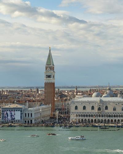 A panoramic view of the Venetian lagoon and skyline, prominently featuring St. Mark’s Campanile (bell tower), the Doge’s Palace, and the waterfront buildings of Piazza San Marco and the Piazzetta, with boats on the water in the foreground.