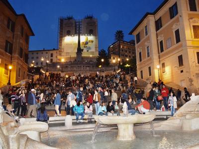 A wide-angle night view of a massive, crowded stone staircase leading up to a church facade covered in scaffolding and a large "MICHAEL KORS" advertisement, with a fountain in the immediate foreground.