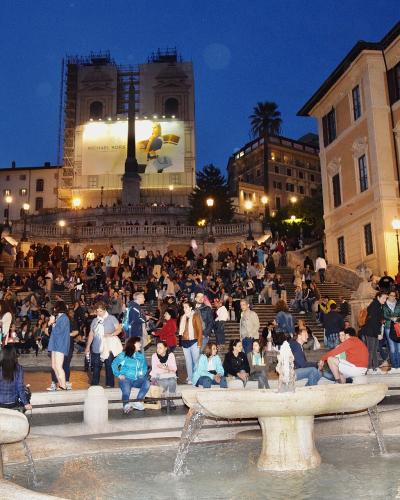 A wide-angle night view of a massive, crowded stone staircase leading up to a church facade covered in scaffolding and a large "MICHAEL KORS" advertisement, with a fountain in the immediate foreground.