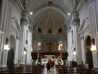 The image shows the interior nave of a large church with white walls, ornate columns, wooden pews, a central aisle, and an altar area featuring a crucifix, organ pipes, and stained-glass windows; people are walking inside, suggesting it is an acti