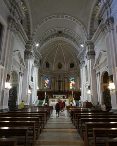 The image shows the interior nave of a large church with white walls, ornate columns, wooden pews, a central aisle, and an altar area featuring a crucifix, organ pipes, and stained-glass windows; people are walking inside, suggesting it is an acti