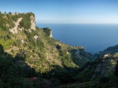 A high-angle landscape view of steep, terraced hillsides covered in lush green vegetation and dotted with small houses, overlooking the deep blue Tyrrhenian Sea under a clear sky.