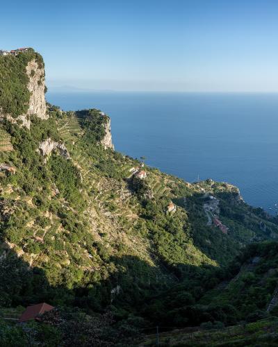 A high-angle landscape view of steep, terraced hillsides covered in lush green vegetation and dotted with small houses, overlooking the deep blue Tyrrhenian Sea under a clear sky.