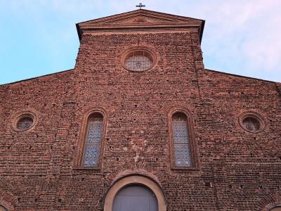A low-angle photograph of a large, red-brick church facade featuring a central pediment topped with a cross, a circular window, two tall arched windows, and smaller circular windows on the sides.