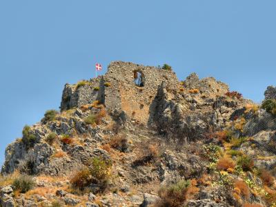 The image shows the stone ruins of a medieval fortress atop a rocky, vegetated hill under a clear blue sky, with a red and white flag (resembling the flag of Genoa or a similar heraldic banner) flying from the highest point of the ruins.