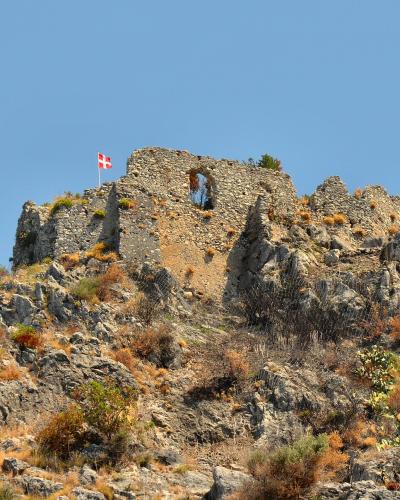 The image shows the stone ruins of a medieval fortress atop a rocky, vegetated hill under a clear blue sky, with a red and white flag (resembling the flag of Genoa or a similar heraldic banner) flying from the highest point of the ruins.