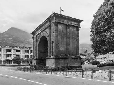 A black-and-white photograph of a large, ancient Roman triumphal arch with a single central archway, flanked by pilasters and topped with a flat entablature and a small flag; it stands in an urban plaza with modern buildings, trees, and mountains