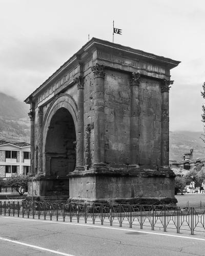 A black-and-white photograph of a large, ancient Roman triumphal arch with a single central archway, flanked by pilasters and topped with a flat entablature and a small flag; it stands in an urban plaza with modern buildings, trees, and mountains