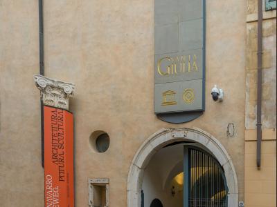 The image shows the exterior entrance of a building with a sign reading "SANTA GIULIA MUSEO CIVICO CITTÀ" and an orange exhibition banner for "JUAN NAVARRO BALDEWEG" at the "Museo di Santa Giulia Brescia".