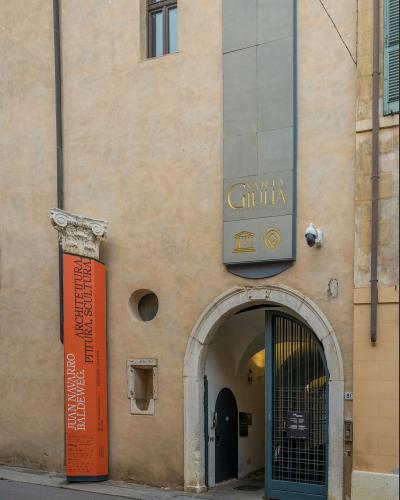 The image shows the exterior entrance of a building with a sign reading "SANTA GIULIA MUSEO CIVICO CITTÀ" and an orange exhibition banner for "JUAN NAVARRO BALDEWEG" at the "Museo di Santa Giulia Brescia".