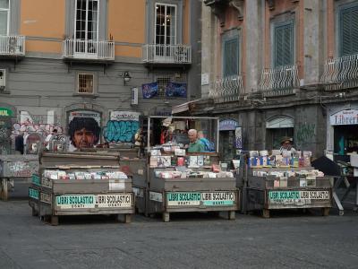 The image shows an outdoor used book market with wooden stalls labeled "LIBRI SCOLASTICI USATI" in a paved urban square, featuring buildings with graffiti, balconies, and a shop sign reading "Librerie" in the background.