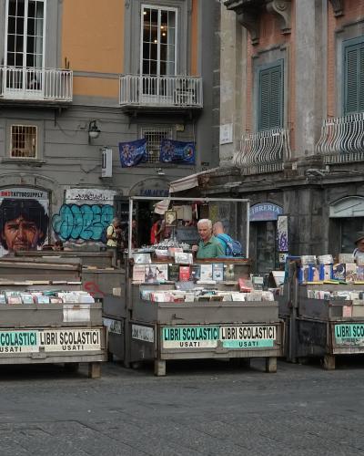 The image shows an outdoor used book market with wooden stalls labeled "LIBRI SCOLASTICI USATI" in a paved urban square, featuring buildings with graffiti, balconies, and a shop sign reading "Librerie" in the background.