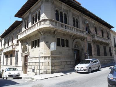 A corner view of a multi-story stone building with arched windows, balconies, and a sign reading "ROSSI" on the facade, situated on a street with parked cars under a clear blue sky.