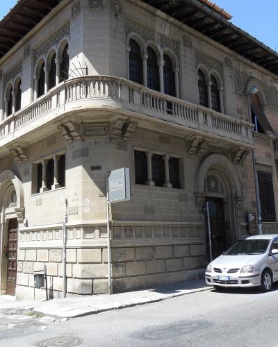 A corner view of a multi-story stone building with arched windows, balconies, and a sign reading "ROSSI" on the facade, situated on a street with parked cars under a clear blue sky.