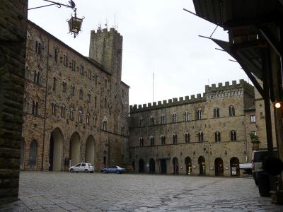 A cobblestone piazza flanked by medieval stone buildings with arched loggias and crenellated towers, including a prominent tower on the left building and a fortified wall on the right; a white car and blue police car are parked near the center, un