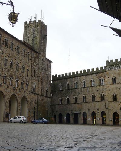 A cobblestone piazza flanked by medieval stone buildings with arched loggias and crenellated towers, including a prominent tower on the left building and a fortified wall on the right; a white car and blue police car are parked near the center, un