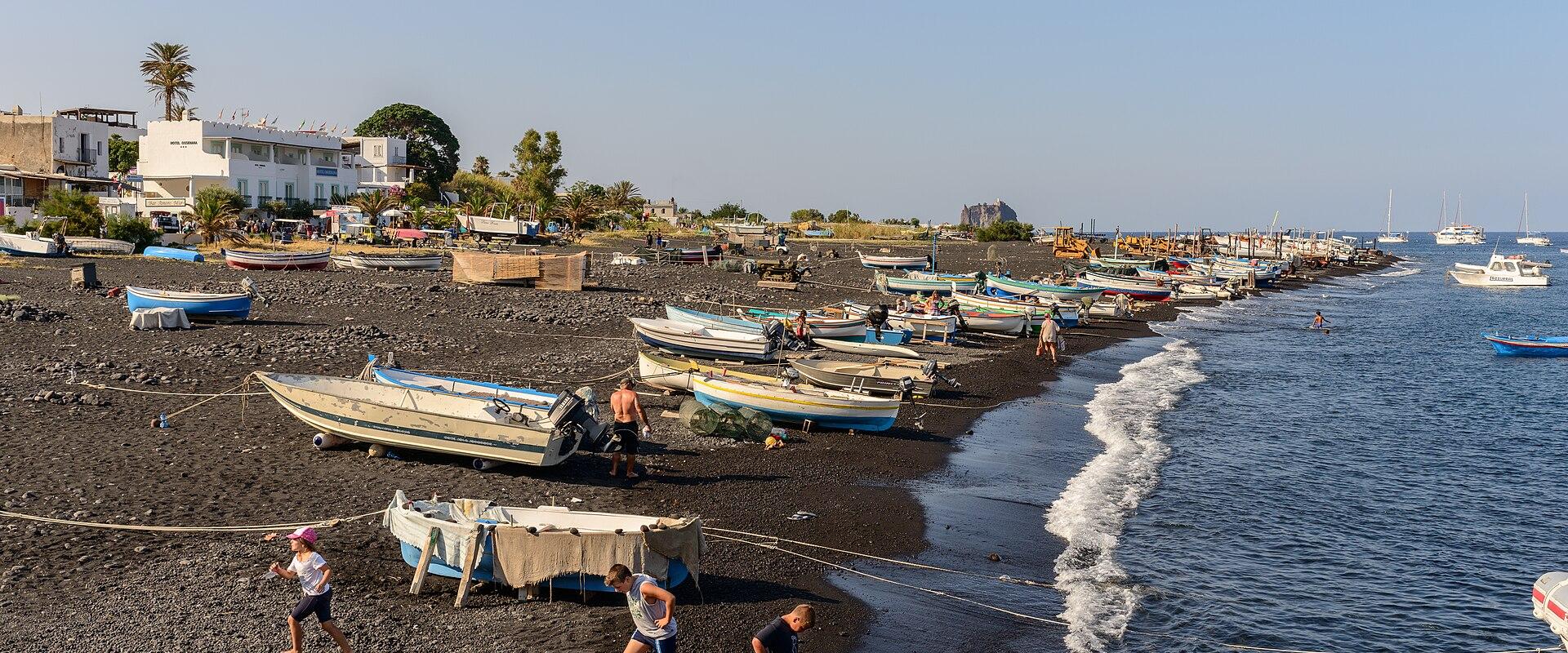 A black volcanic pebble beach lined with numerous small fishing boats pulled ashore, with people walking near the water’s edge and buildings including a white hotel-like structure in the background under a clear blue sky.